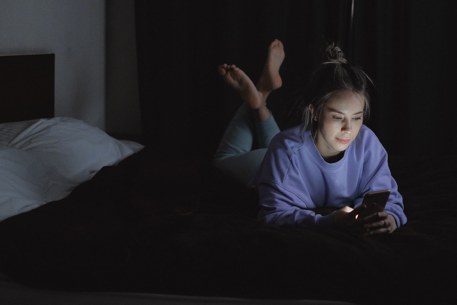A young woman lying in bed at night, her face lit by the glow of her phone as she scrolls