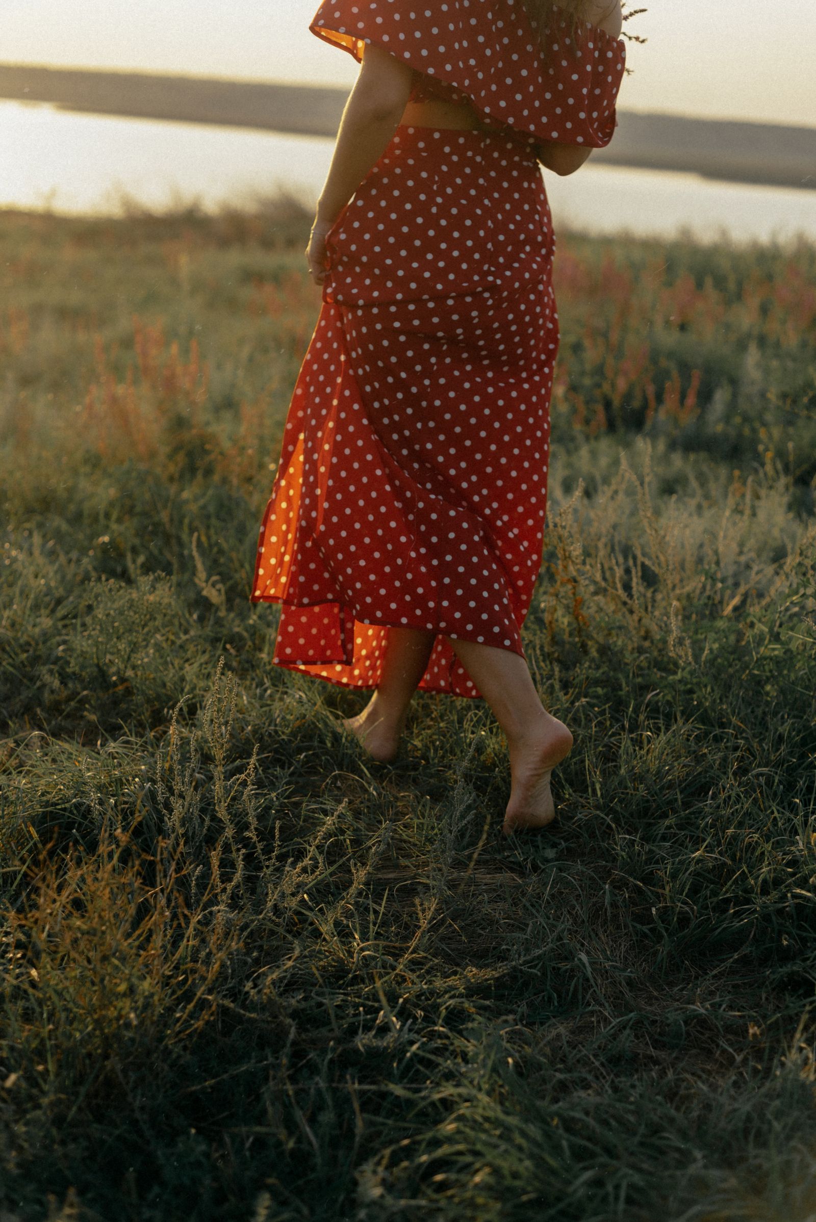 A woman in a red dress walking barefoot through a sunlit meadow at sunrise